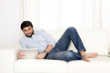 Genç çekici İspanyol adam evde beyaz kanepede dijital tablet veya pad kullanarak