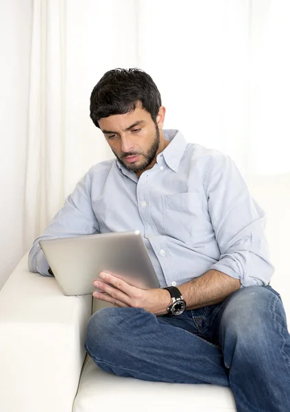 Jovem atraente homem hispânico em casa no sofá branco usando tablet digital ou pad — Fotografia de Stock