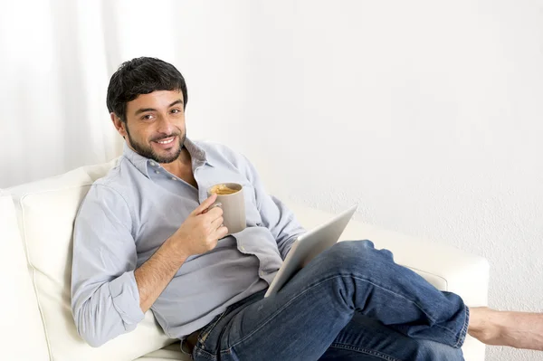 Joven y atractivo hombre hispano en casa en un sofá blanco usando tableta digital o almohadilla —  Fotos de Stock