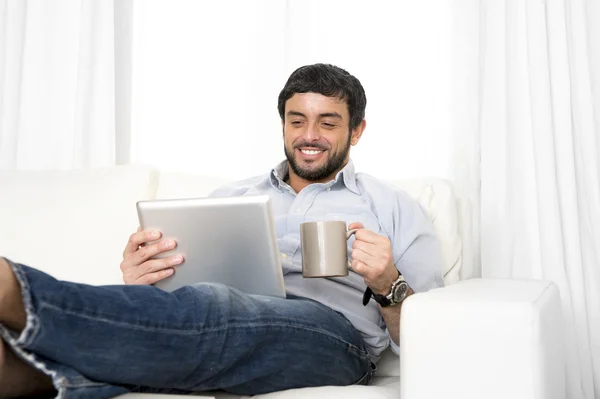 Jovem atraente homem hispânico em casa no sofá branco usando tablet digital ou pad — Fotografia de Stock