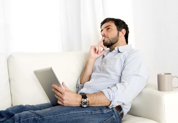 Joven y atractivo hombre hispano en casa en un sofá blanco usando tableta digital o almohadilla —  Fotos de Stock
