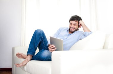 Genç çekici İspanyol adam evde beyaz kanepede dijital tablet veya pad kullanarak