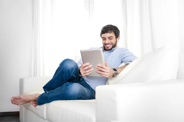 Vonzó hispán fiatalember otthon fehér kanapén, digitális tabletta vagy pad segítségével — Stock Fotó