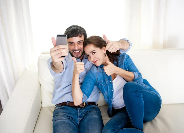 Nice attractive young couple sitting together in sofa couch taking selfie photo with mobile phone — Stock Photo, Image