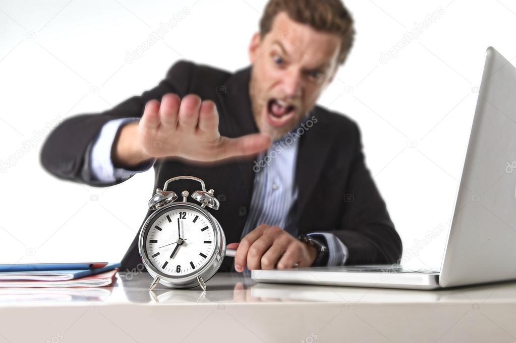 Exploited businessman at office desk stressed and frustrated with  alarm clock in out of time and deadline concept