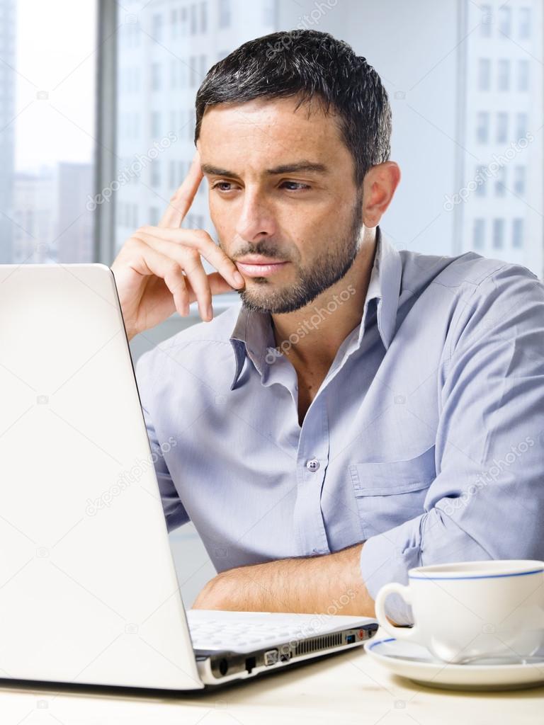 attractive businessman working on computer at office desk in front of skyscraper window