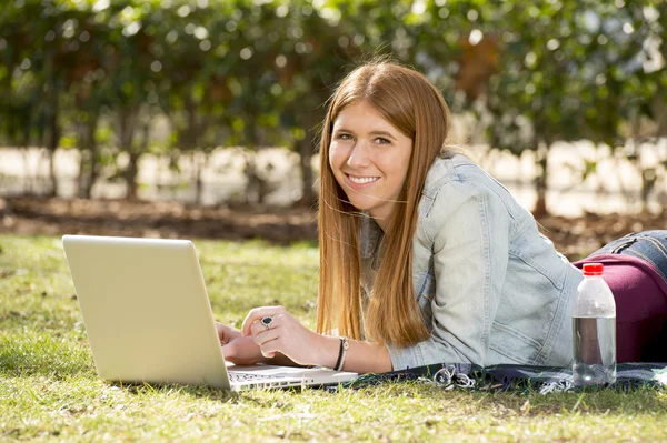 Молодая студентка лежит на траве в парке с компьютером изучения или серфинга в Интернете — стоковое фото