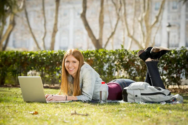Молодая студентка лежит на траве в парке с компьютером изучения или серфинга в Интернете — стоковое фото