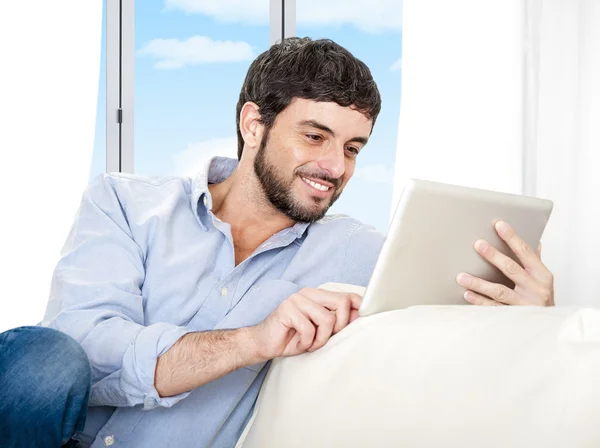 Joven y atractivo hombre hispano en casa sentado en un sofá blanco usando una tableta digital — Foto de Stock