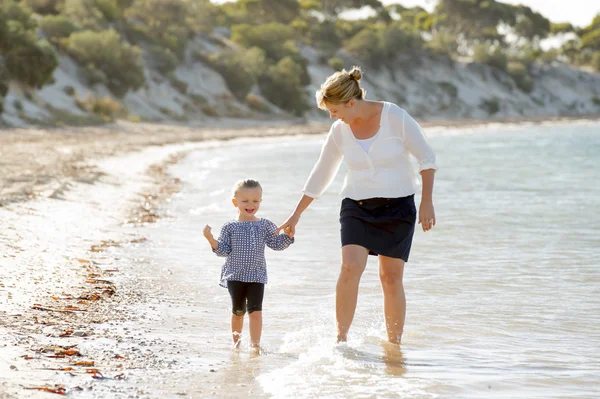 Happy mother holding hand of sweet blond little daughter walking together on sand at beach sea shore — Stock Photo, Image