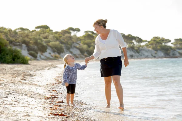 Happy mother holding hand of sweet blond little daughter walking together on sand at beach sea shore — Stock Photo, Image