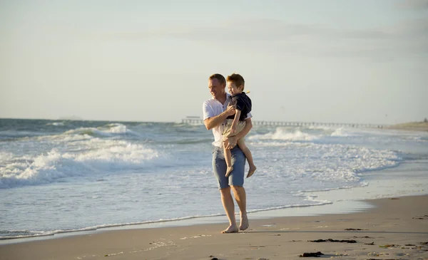 Молодой счастливый отец держит в руках маленького сына гуляющего по пляжному песку перед морскими волнами — стоковое фото