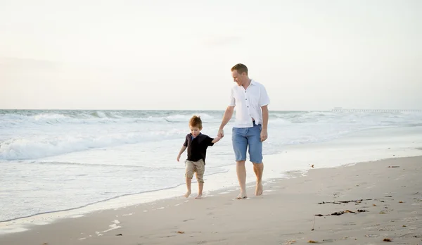 Счастливый отец держит за руку маленького сына, идущего вместе по пляжу босиком — стоковое фото