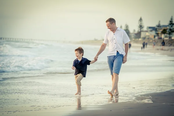 Счастливый отец держит за руку маленького сына, идущего вместе по пляжу босиком — стоковое фото