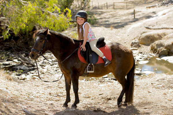 sweet young girl 7 or 8 years old riding pony horse smiling happy wearing safety jockey helmet in summer holiday