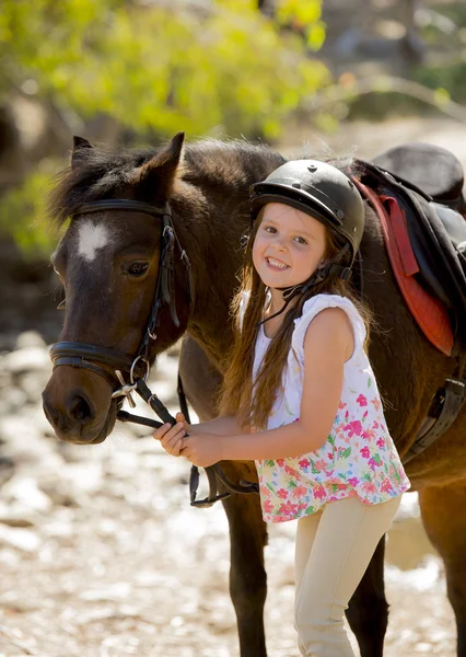 Νεαρό κορίτσι 7 ή 8 ετών εκμετάλλευση χαλινάρι μικρό πόνυ αλόγου, χαμογελά ευτυχισμένος φορώντας jockey κράνους ασφαλείας σε καλοκαιρινές διακοπές — Φωτογραφία Αρχείου