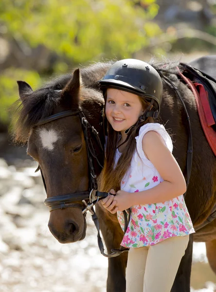 Jovem menina 7 ou 8 anos de idade segurando freio de cavalo pequeno pônei sorrindo feliz vestindo capacete de segurança jockey em férias de verão — Fotografia de Stock