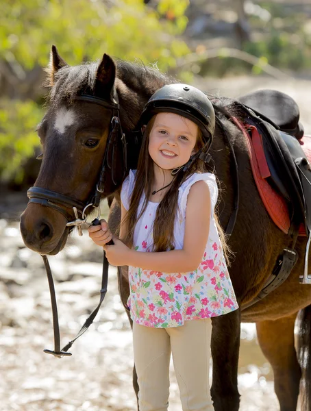 Jovem menina 7 ou 8 anos de idade segurando freio de cavalo pequeno pônei sorrindo feliz vestindo capacete de segurança jockey em férias de verão — Fotografia de Stock
