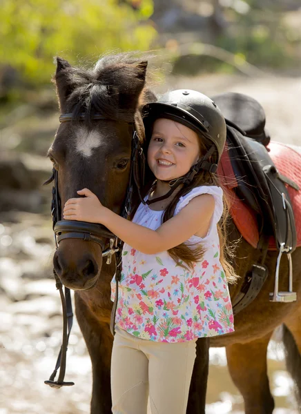 Γλυκό όμορφη νεαρή κοπέλα 7 ή 8 ετών αγκαλιάζει το κεφάλι του αλόγου μικρό πόνυ, χαμογελά ευτυχισμένος φορώντας κράνος ασφαλείας αναβάτης σε καλοκαιρινές διακοπές — Φωτογραφία Αρχείου