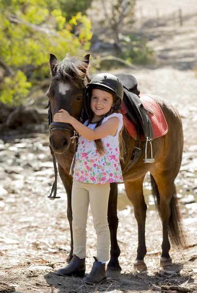 sweet beautiful young girl 7 or 8 years old hugging head of little pony horse smiling happy wearing safety jockey helmet in summer holiday