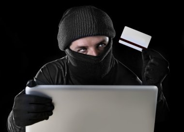 man in black holding credit card using computer laptop for criminal activity hacking password and private information clipart