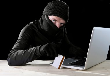 man in black holding credit card using computer laptop for criminal activity hacking password and private information clipart