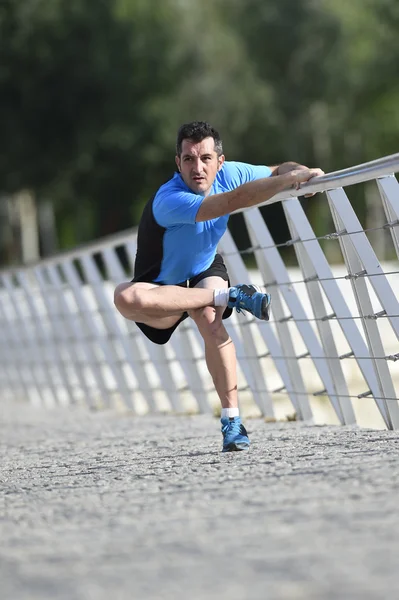 Lekkoatletka mężczyzna rozciąganie nogi rozgrzanie mięśni przed uruchomieniem treningu opierając się na poręczy miasto miejski park — Zdjęcie stockowe