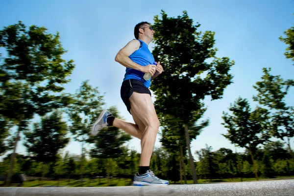 Young man with athletic runner legs holding isotonic energy drink while running in city park — Stock Photo, Image