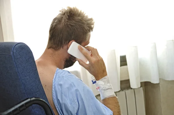 Jonge man bij ziekenhuis kamer dragen van patiënt pyjama's zitten bij het raam praten op mobiele telefoon — Stockfoto