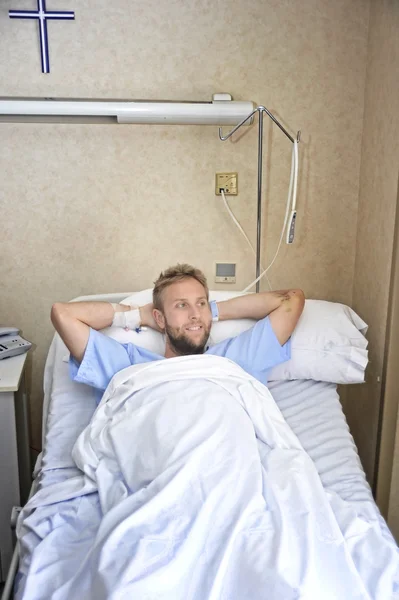 Young American man lying in bed at hospital room sick or ill but feeling relaxed  smiling happy and positive — Stock Photo, Image