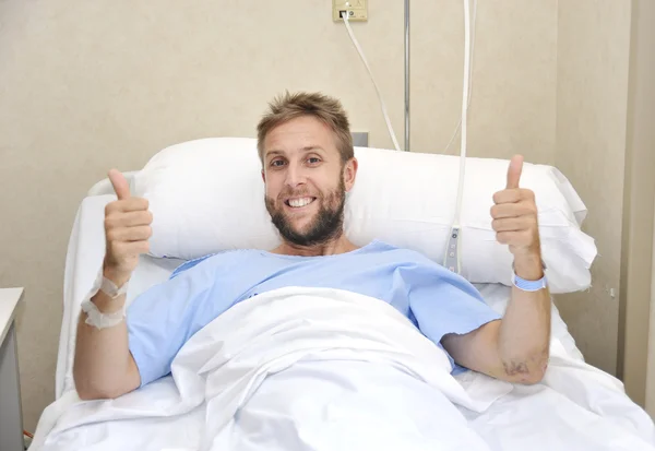 Young American man lying in bed at hospital room sick or ill but giving thumbs up smiling happy and positive — Zdjęcie stockowe