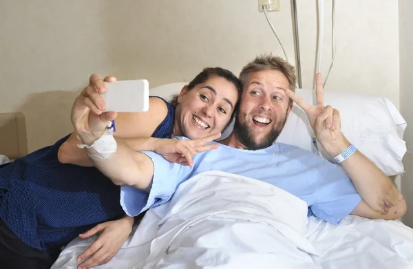 Young couple taking selfie photo at hospital room with man lying in clinic bed — Stock Photo, Image