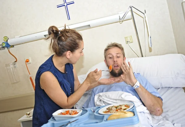 Młoda żona stara się karmić jego mąż niechętnie leżąc w łóżku w szpitalnej sali chorych po wypadku — Zdjęcie stockowe