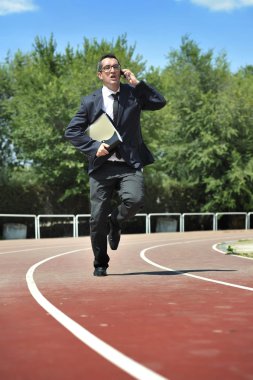 Businessman in suit and necktie carrying folder portfolio and files running in stress on athletic track talking on mobile phone looking tired and stressed