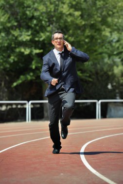 Businessman in suit and necktie running in stress on athletic track talking on mobile phone looking tired overworked and stressed in work urgent problems concept