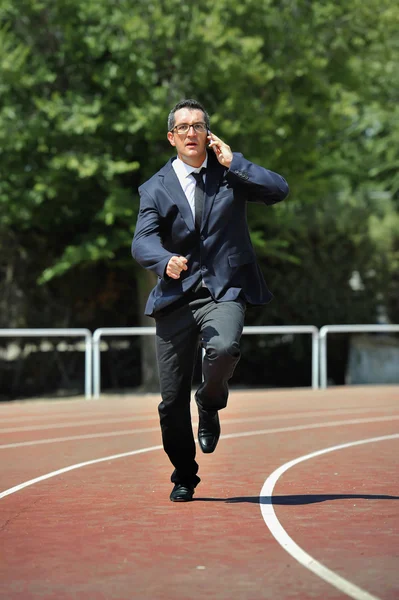 Geschäftsmann in Anzug und Krawatte läuft im Stress auf der Leichtathletikbahn, telefoniert mit dem Handy und wirkt müde, überarbeitet und gestresst in der Arbeit. — Stockfoto