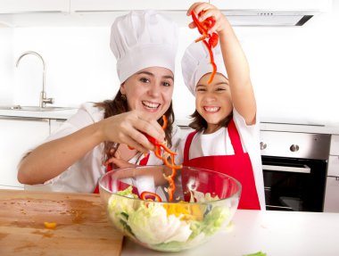 happy mother and little daughter at home kitchen preparing paprika salad in apron and cook hat