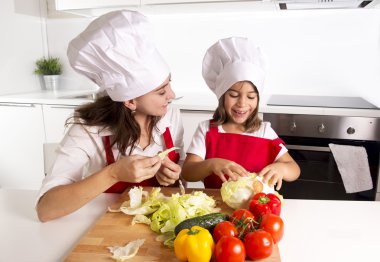 happy mother and little daughter at home kitchen preparing salad in apron and cook hat