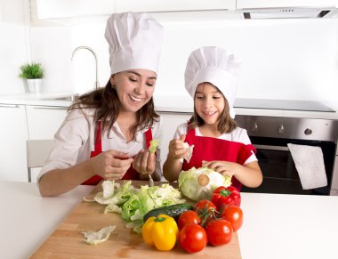 happy mother and little daughter at home kitchen preparing salad in apron and cook hat