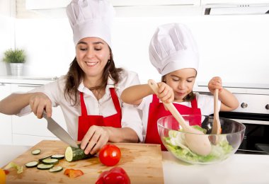 young mother and little daughter at house kitchen preparing salad for lunch wearing apron and cook hat