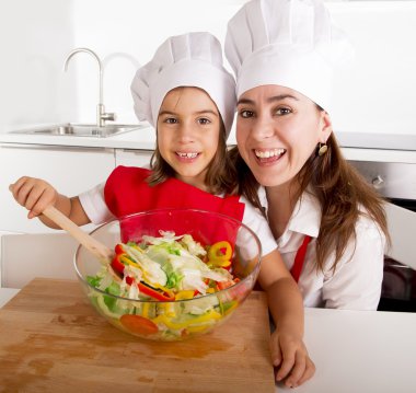 happy mother and little daughter at home kitchen preparing salad in apron and cook hat