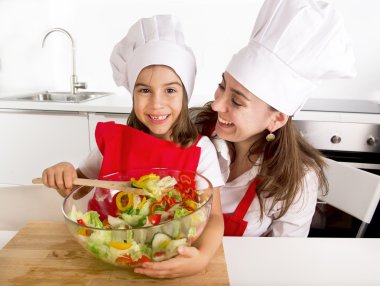 happy mother and little daughter at home kitchen preparing salad in apron and cook hat