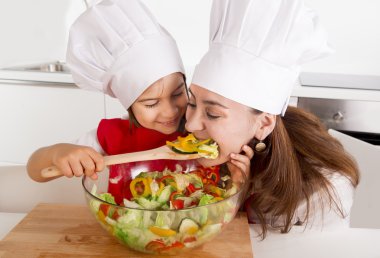 happy mother and little daughter at home kitchen preparing salad in apron and cook hat