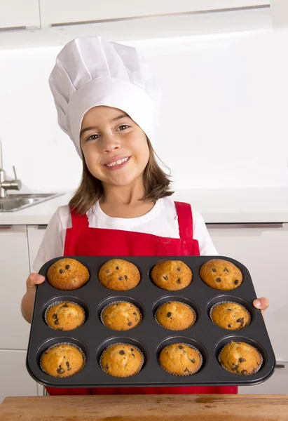 Proud female child presenting her self made muffin cakes learning baking wearing red apron and cook hat smiling happy — Stock Photo, Image