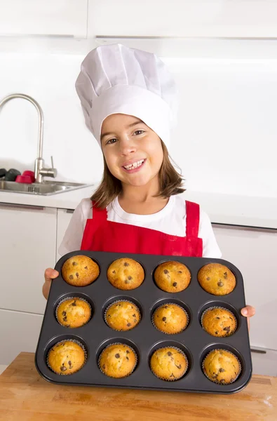 Proud female child presenting her self made muffin cakes learning baking wearing red apron and cook hat smiling happy — Stock Photo, Image