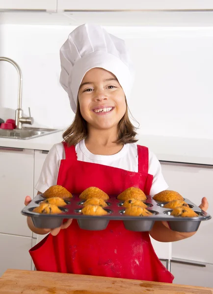 Proud female child presenting her self made muffin cakes learning baking wearing red apron and cook hat smiling happy — Stock Photo, Image