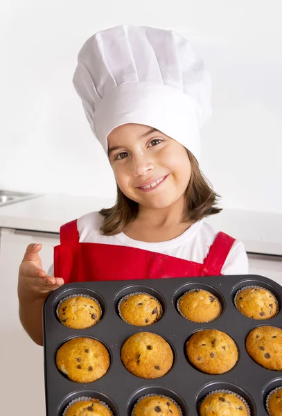 Proud female child presenting her self made muffin cakes learning baking wearing red apron and cook hat smiling happy — Stock Photo, Image