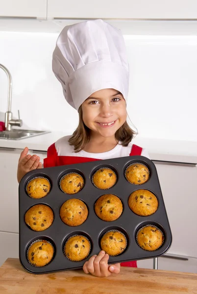 Proud female child presenting her self made muffin cakes learning baking wearing red apron and cook hat smiling happy — Stock Photo, Image