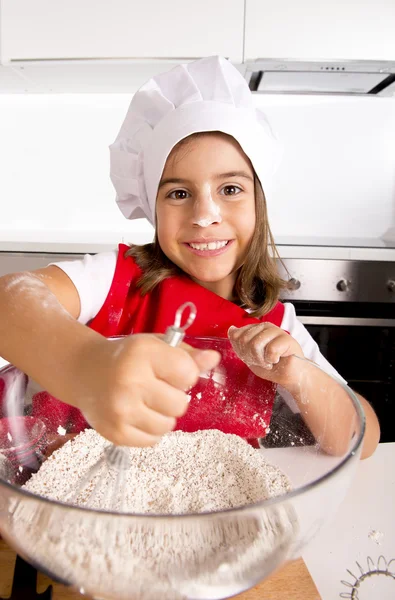 Glückliches kleines Mädchen lernt, Mehl in Schüssel zu backen und trägt rote Schürze und Kochmütze und lächelt zufrieden — Stockfoto