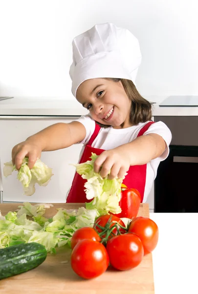 Happy little girl playing with vegetables at home kitchen in apron and cook hat — Stock Photo, Image